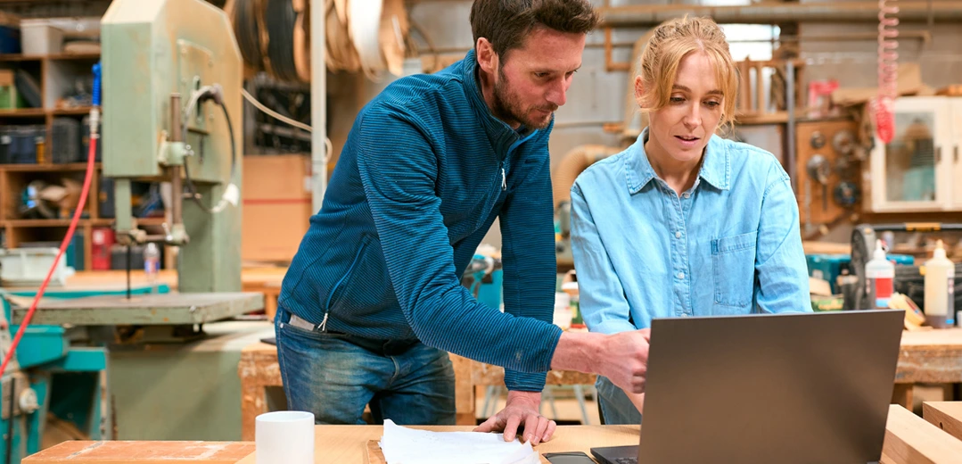 A man and a woman, standing in a workshop, discussing their business needs while looking at a laptop.