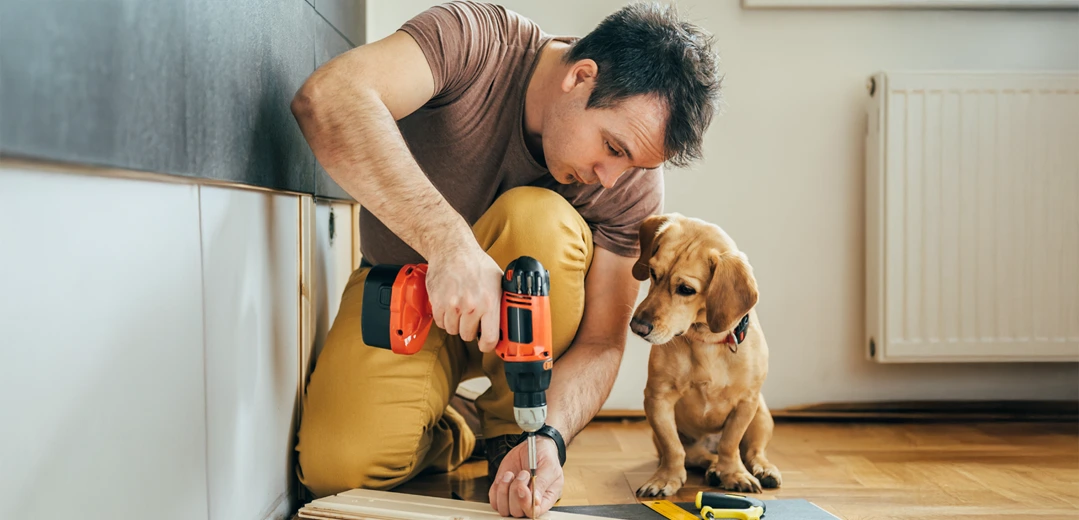 A man using a drill to make home improvements with his dog looking on.