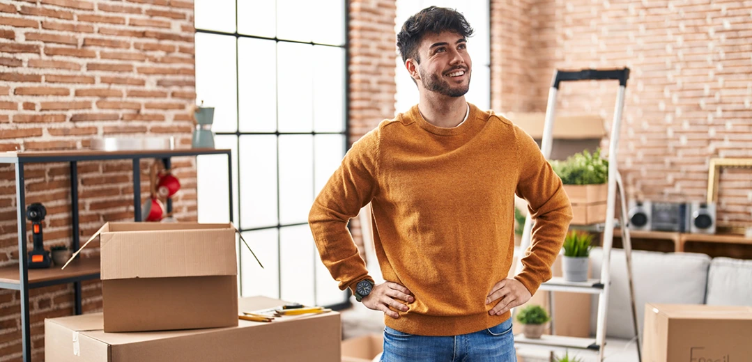 Smiling young adult man standing in his new apartment surrounded by moving boxes.