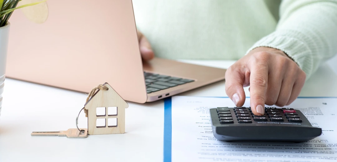 Image of a person using a calculator and a laptop to compare mortgage closing costs. A house keychain is on the desk.